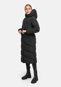 Long black puffer coat with a hood, featuring diagonal quilted patterns, snap buttons, and side pockets. Worn over black leggings and boots.