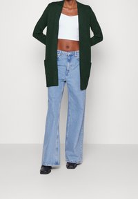 Zielony kardigan z otwartym przodem, długimi rękawami i dwiema przednimi kieszeniami, w połączeniu z jasnoniebieskimi spodniami o szerokich nogawkach i czarnymi butami.