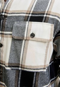 Karirana košulja s mješavinom bijele, crne i tamno smeđe boje. Napravljena od mekog materijala, ima dugmad na prednjem dijelu i džep na prsima s kopčanjem na dugme.