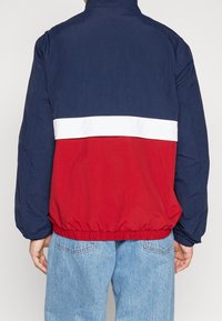 Kurtka z blokami kolorów, z granatową górą, czerwoną dolną częścią i białym paskiem pomiędzy nimi. Wykonana z lekkiego materiału z elastycznymi mankietami.