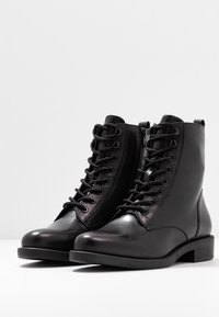 Las botas de tobillo de cuero negro presentan una punta redonda, cordones en la parte frontal, cremallera lateral y un tacón bajo y cuadrado. Textura suave con detalles mínimos.