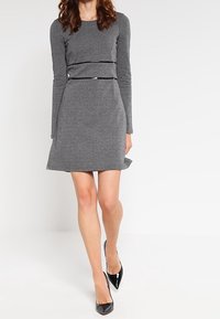 Frau, die ein graues, langärmliges, strukturiertes Strickkleid mit schwarzem Paspel und schwarzen spitzen Absatzschuhen trägt, steht vor einem schlichten weißen Hintergrund.