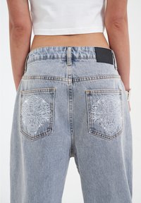 Jeans de mezclilla azul claro con cortes holgados, que cuentan con bolsillos traseros adornados con intrincados bordados de tigre blanco y un parche de cuero negro.