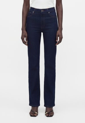 Kvinde iført mørkeblå jeans med lige ben, hvid top og sorte sandaler med gulddetaljer, står foran en ensfarvet baggrund.