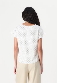 Weiße Bluse mit kurzen Ärmeln, schwarzen Punkten, Rundhalsausschnitt und Rüschensäumen; leichtes Material und lässiger Schnitt.