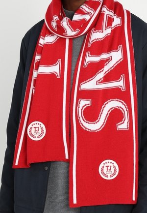 Écharpe rouge avec de grandes lettres blanches "TJ" et des badges décoratifs, portée sur un pull gris et une veste sombre.