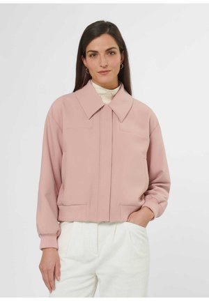 Frau trägt eine hellrosa Jacke mit weitem Kragen über einem weißen Rollkragenpullover, kombiniert mit hoch taillierten weißen Hosen, steht mit einer Hand in der Tasche.