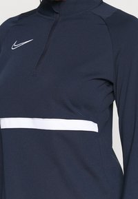 Tmavě modrý dlouhý rukáv s půlzipem, vybavený bílým vodorovným pruhem a logem Nike na hrudi. Hladká textura látky.