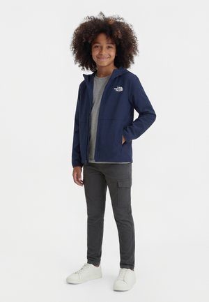 Enfant aux cheveux bouclés portant une veste à fermeture éclair bleu marine, un pantalon cargo gris et des baskets blanches, debout avec une main dans la poche.