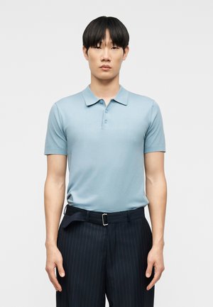 Jeune homme aux cheveux noirs et raides portant un polo bleu clair à manches courtes et un pantalon sombre à fines rayures, debout devant un fond blanc uni.