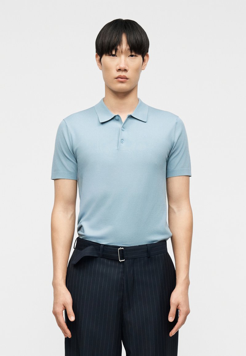 Νέος άνδρας με ίσια μαύρα μαλλιά φορεί ανοιχτό μπλε κοντομάνικο πόλο πουκάμισο και σκούρο παντελόνι με λεπτές ρίγες, στέκεται μπροστά από έναν απλό λευκό φόντο.