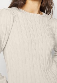 Světlý béžový svetr s texturovaným vzorem kabelového pletení a žebrovaným výstřihem. Detailní pohled zdůrazňující měkký materiál a design.