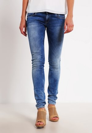 Femme portant un jean skinny bleu délavé et des sandales à talons beiges perforées à bout ouvert, debout devant un fond blanc.