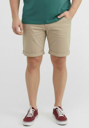 Mann trägt beige hochgekrempelte Shorts, grünes Hemd und rote Schnürsneaker und steht mit einer Hand in der Tasche.