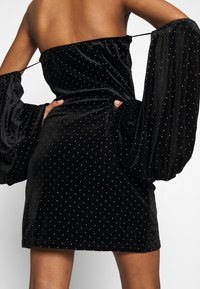 Mini-robe noire en velours sans bretelles avec des pois dorés et des manches longues volumineuses dévoilant les épaules, portée par une personne avec une main sur la hanche.