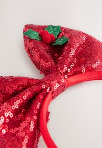 Rotes Pailletten-Stirnband mit großer Schleife und grünem Stechpalmen-Akzent mit roten Bommel. Glatte Textur mit glänzenden, reflektierenden Pailletten.