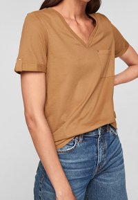 Brun V-ringad t-shirt med rullade ärmar, utrustad med en liten bröstficka och en mjuk bomullstextur. Bärs tillsammans med blå denimjeans.