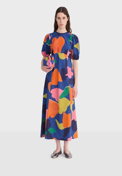 Femme portant une robe longue bleue avec de grands motifs floraux colorés et des manches bouffantes, tenant une pochette assortie au motif.