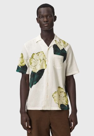Ung mand iført en hvid kortærmet skjorte med store grønne og gule blomstermønstre og brune bukser.