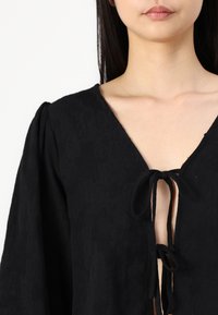 Blusa nera con tessuto testurizzato, scollatura a V profonda con lacci e maniche a sbuffo. Il design presenta motivi sottili.