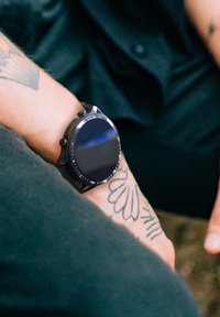 Orologio smartwatch nero con cassa rotonda, design minimalista, superficie in vetro liscia, indossato su un polso decorato con tatuaggi floreali.