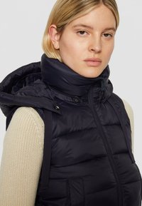 Granatowa pikowana kamizelka puchowa z wysokim kołnierzem i kapturem. Posiada zamek błyskawiczny oraz boczne kieszenie. Noszona na kremowym swetrze o fakturowanym splocie.