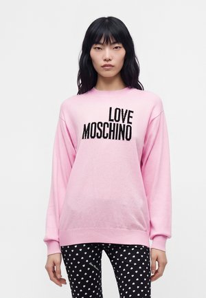 Жена с дълга черна коса носи розов пуловер с текст "LOVE MOSCHINO" и черни клинове с бели сърчица.
