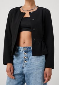 Veste péplum noire avec encolure ronde, boutons pression à l'avant et manches longues. Portée sur un crop top noir et un jean en denim bleu taille haute.