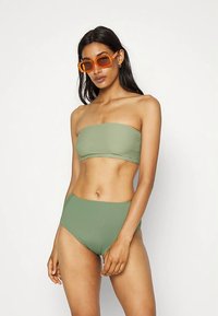 Top de biquíni verde estilo bandeau, com textura canelada, combinado com cuecas de biquíni verdes de cintura alta. Modelo a usar óculos de sol laranja.