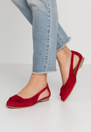Stopy noszące czerwone zamszowe baleriny typu slingback z ozdobną kokardką połączone z jasnoniebieskimi jeansami z postrzępionym brzegiem na białym tle podłogi.