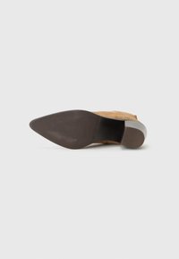 Vue de dessous d'une chaussure à talon haut marron avec un bout pointu et un talon carré, sur fond gris clair.
