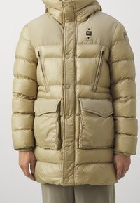 Beżowa pikowana kurtka o połyskującym wykończeniu, wyposażona w kaptur, zatrzaski, dwie duże przednie kieszenie oraz zapinane na zamek kieszenie na piersi.