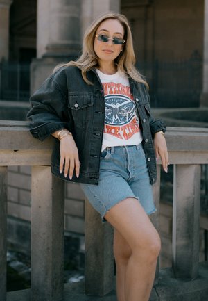 Svart denimjacka, vit grafisk t-shirt med blå och orange design, ljusblå denimshorts och solglasögon. Avslappnad, ledig passform.