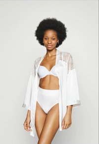 Afro soenguga naine, kes kannab valget rinnahoidjat, kõrge vöökohtadega aluspükse ja läbipaistvat pitsist satiinist kimono, seistes lihtsa heledal taustal.
