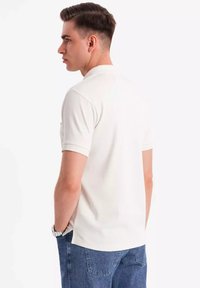 Hvid polo t-shirt til mænd med klassisk krave, korte ærmer og lige skæring. Bæres med blå denim jeans, vist fra bagview.