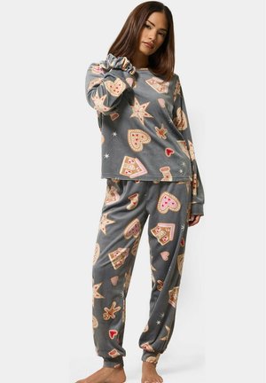 Femme portant un ensemble de pyjama gris décoré de maisons en pain d'épices, étoiles, cœurs, bas et bonshommes en pain d'épices, debout pieds nus.