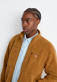 Young man with braided hair wears light blue button-up and brown fleece jacket, looking slightly to the side against a plain white background.