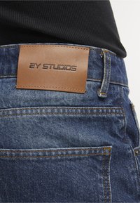 Pochette en cuir marron avec l'étiquette "2Y STUDIOS" sur la ceinture d'un jean en denim bleu porté avec un haut noir.