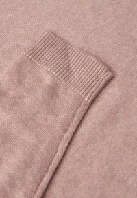 Hellrosa gestrickter Pulloverstoff mit einem gerippten Bündchen. Weiche Textur und subtile Farbnuancen. Nahaufnahme zeigt Stoffdetail und Naht.