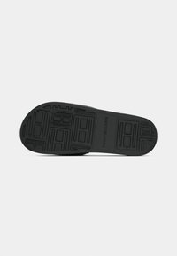 Semelle de chaussure en caoutchouc noir avec un motif de crampons géométrique et le logo "Tommy Hilfiger" embossé en son centre.