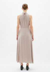 Langes, ärmelloses, hellbeiges Kleid mit gerafftem Ausschnitt und Schlüssellochdetail am Rücken, das bis zum Saum unterhalb der Knöchel reicht; getragen mit schwarzen High Heels.
