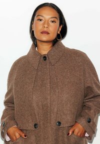 Manteau en laine marron avec un col montant, une silhouette ample et des boutons noirs. Il dispose de deux poches latérales et d'une finition texturée.