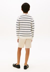 Παιδί που στέκεται με την πλάτη, φορώντας λευκό πουκάμισο με μακριά μανίκια και ναυτιλιακές ρίγες, μπεζ σορτς και μαύρα μοκασίνια σε λευκό φόντο.