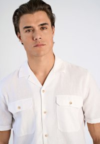 Camicia bianca a maniche corte realizzata in tessuto leggero, con due tasche applicate, chiusure a bottoni e colletto a punta.