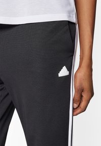 Svarta sportbyxor i ett slätt tyg med vita sidostråk och en liten vit logga. Inkluderar fickor och en figurnära design.