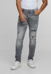 Egy férfi elhasználódott szürke, festékfoltos farmert, fehér sportcipőt és fehér ujjatlan pólót visel, egyik kezével a zsebében áll.