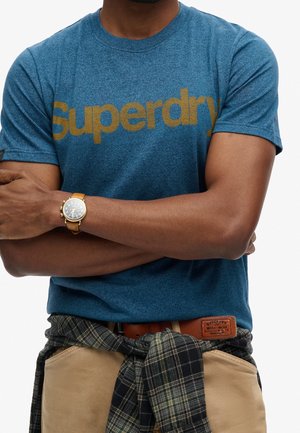 Homme portant un t-shirt bleu Superdry, un pantalon beige, une chemise à carreaux nouée autour de la taille, et une montre-bracelet avec un bracelet en cuir marron.