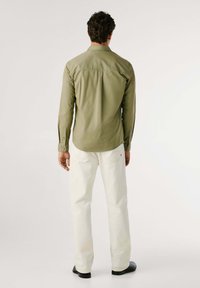 Camicia a maniche lunghe di un verde oliva chiaro con una superficie liscia, polsini abbottonati e orlo curvo, abbinata a pantaloni color crema e scarpe nere.