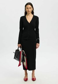 Set di cardigan e gonna in maglia a costine nera, con bottoni sul davanti. La modella tiene una borsa nera con una sciarpa rossa, crema e nera. Scarpe ballerine rosse.