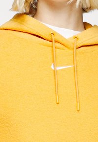 Keltainen puuvillainen huppari, jossa on teksturoitu pinta, etutasku, kiristysnauhallinen huppu ja valkoinen Nike-logo rinnassa.